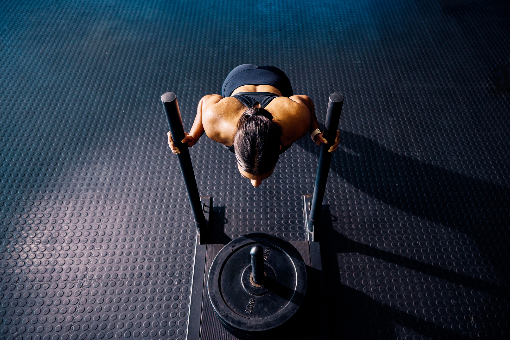 Female athlete pushing a weighted sled in a gym. Top view of a woman performing strength training exercise on a black gym floor. Hyrox Training Concept. Hyrox